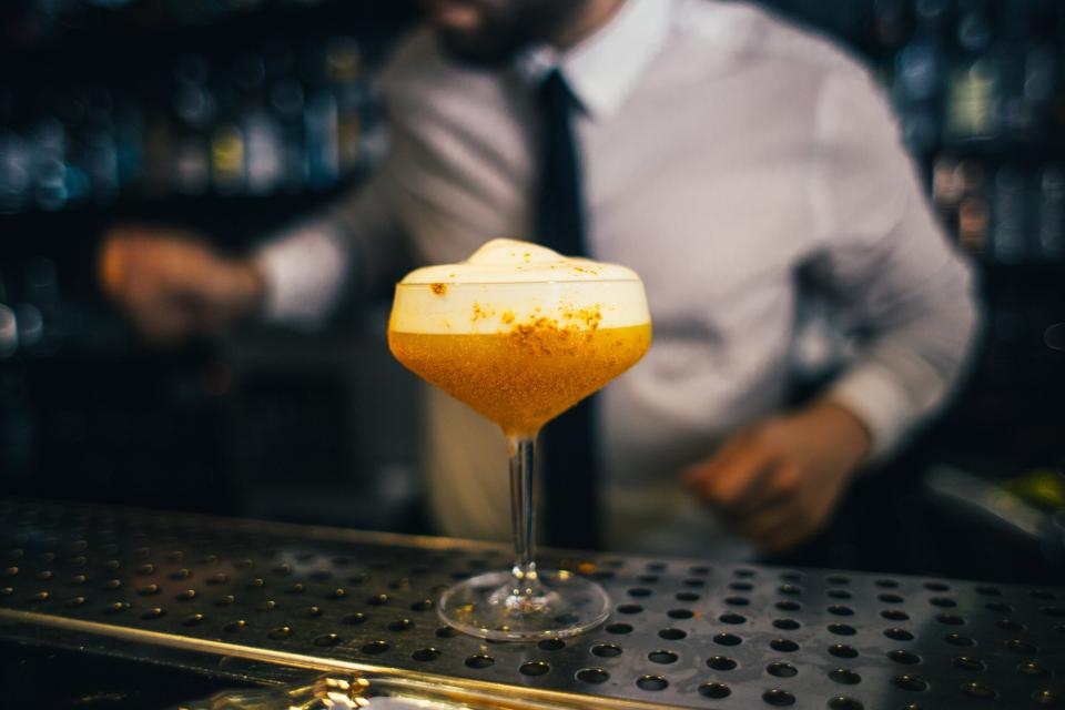 Copa de cóctel sobre la barra metálica de un bar, con el camarero trabajando desenfocado al fondo.