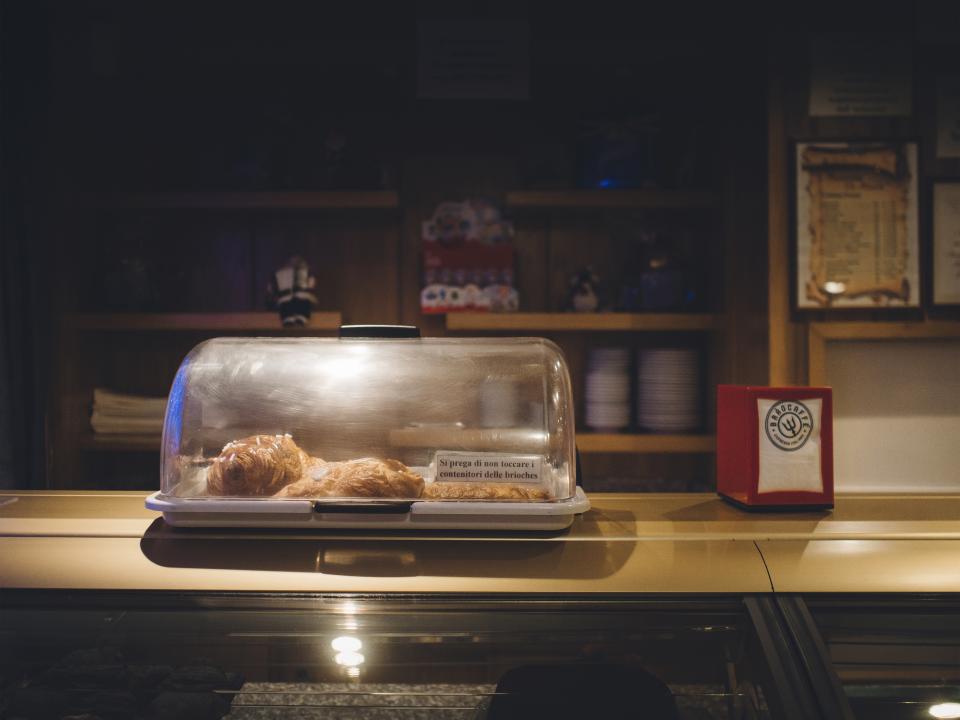 Mostrador de cafetería con una campana de cristal cubriendo unos cruasanes y la estantería de tazas al fondo.