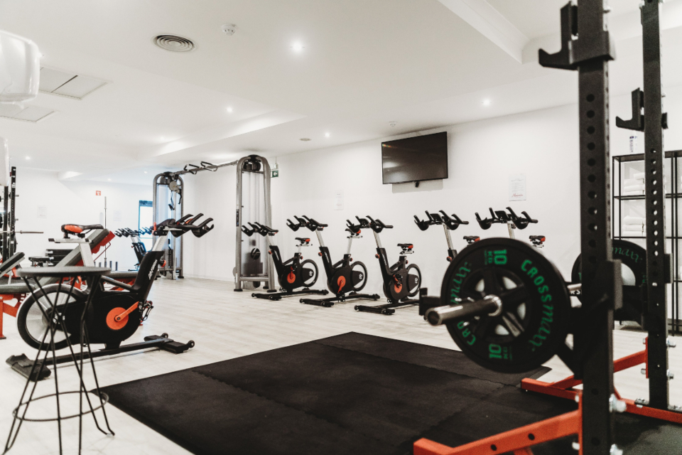 Sala de gimnasio con bicicletas de spinning alineadas y una jaula de pesas con discos en primer plano.