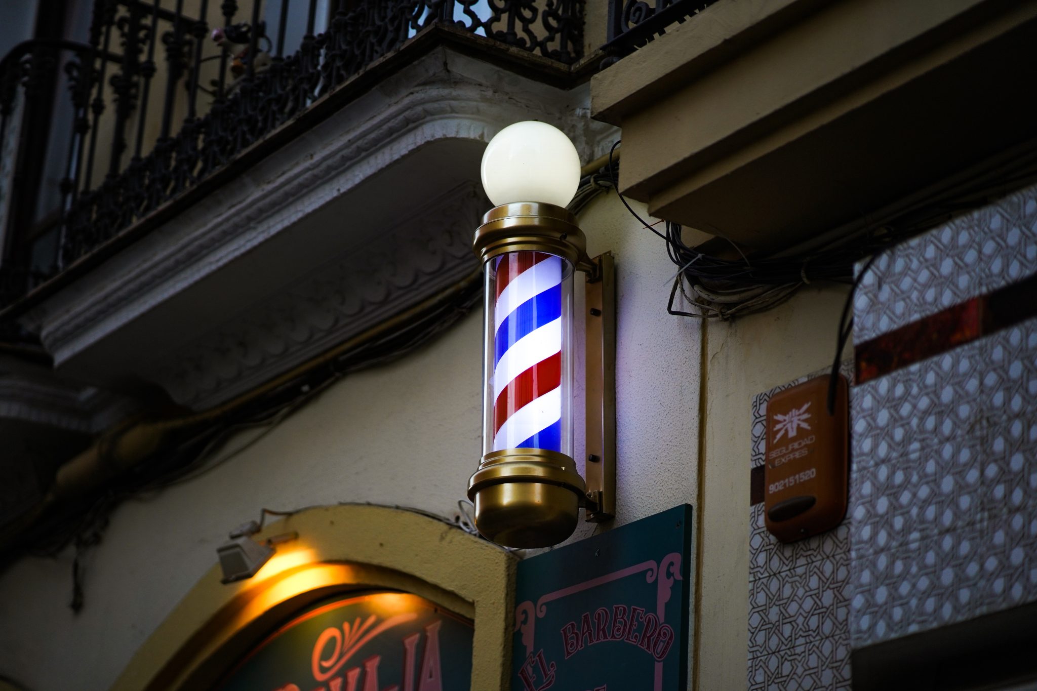 Poste giratorio de barbería iluminado en la fachada de un local, junto al rótulo del establecimiento.