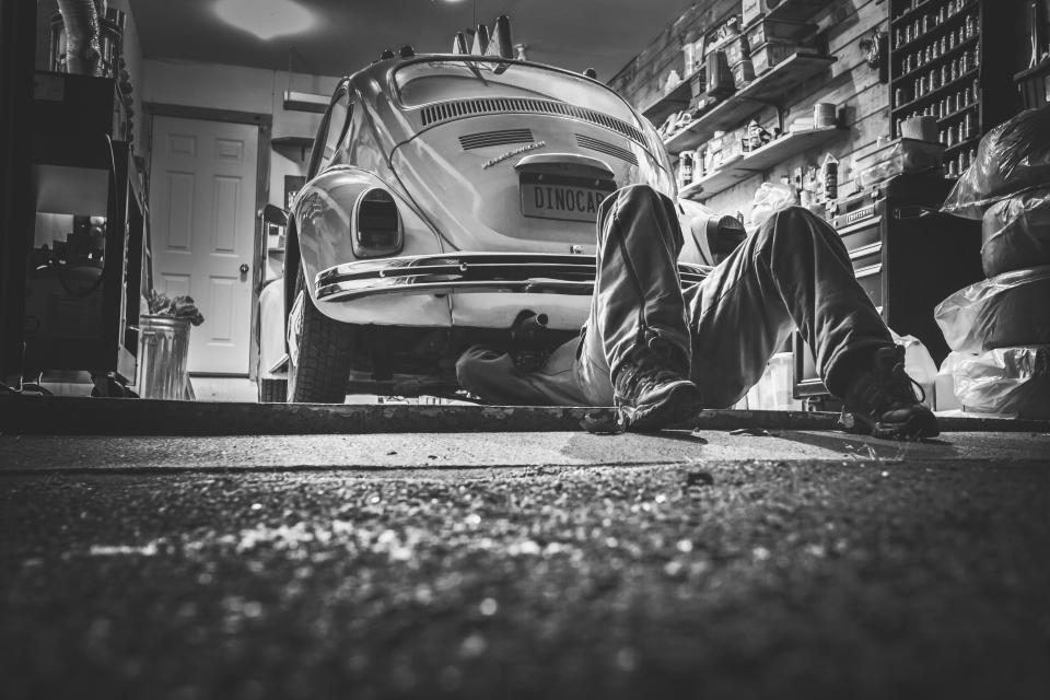 Mecánico tumbado bajo la parte trasera de un Volkswagen Escarabajo en un taller, en blanco y negro.